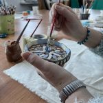 Osoba maluje małą ceramikę w oryginalny wzór, używając pędzla. Kolory jasnobrązowy, niebieski i biały dominują na tle drewnianego stołu. W tle widoczne są przybory malarskie oraz półki.