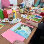 Grupa osób maluje obrazy przy stole, używając jasnych farb i pędzli. Tło to jasna ściana i drewniana szafa. Atmosfera jest kreatywna i radosna.