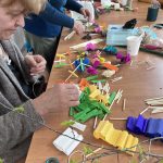 Starsza kobieta tworzy dekoracje z kolorowej krepiny na drewnianym stole. Wokół leżą zielone gałązki, nożyczki i inne materiały. W tle widoczne są kartony i osoby pomagające.