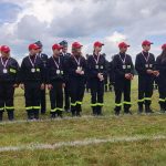 Na zdjęciu grupa młodzieży w czarnych mundurach z medaliami, stojąca na trawie. Noszą czerwone czapki i mają odblaski. W tle jest pochmurne niebo.