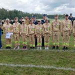 Grupa młodych ludzi w beżowych strojach, z medalami, stoi na trawie. W tle widać drzewa i niebo z chmurami. Obok nich stoi mężczyzna w niebieskiej kurtce, trzymający puchar.