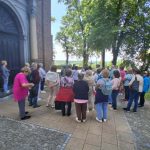 Na zdjęciu grupa osób stoi przed kościołem z cegieł, w towarzystwie zieleni. Tło tworzą drzewa, a widać też drewniany krzyż. Na pierwszym planie kwiaty w wazonie.