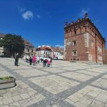 Zdjęcie przedstawia brukowany rynek z ceglanym budynkiem po prawej stronie. Niebieskie niebo i kilka białych chmur w tle. Ludzie spacerują po placu otoczonym kamienicami.