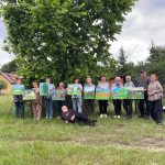 Grupa ludzi stoi na trawie przed drzewem z obrazami na sztalugach. Obrazy są kolorowe, dominują na nich zielone krajobrazy. W tle widać chmury i domki.