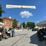 Brukowany rynek z czerwonym gotyckim budynkiem w tle, błękitne niebo z białą chmurką. Banner na festiwal winny, stoliki po prawej.