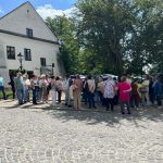 Grupa ludzi stoi na ulicy obok białego budynku. W tle drzewa i zaparkowane samochody. Na niebie widać chmury i słońce.