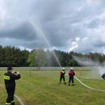 Strażacy w mundurach ćwiczą na zielonym polu, polewając wodę z węży. Tło stanowią drzewa i pochmurne niebo. Kilka osób robi zdjęcia z boku.