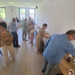 image-1006886228714699 Grupa ludzi na spotkaniu w jasnym, przestronnym pomieszczeniu. Są stoły z jedzeniem, a tło stanowią duże okna i biały krzyż. Sala ma białe ściany i podłogę w jasnym odcieniu drewna.