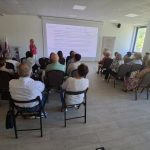 image-2061875141880959 W sali konferencyjnej odbywa się prezentacja. Kobieta w różowej bluzce stoi przy białym ekranie. Uczestnicy siedzą na krzesłach, a światło wpada przez duże okna.