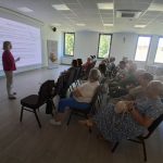 image-4500924073475616 Grupa osób siedzi w sali konferencyjnej, słuchając prezentacji. Kobieta w różowej bluzce stoi przed ekranem. Wnętrze jest jasne, z widokiem na drzewa za oknem.