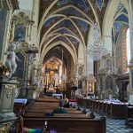 Wnętrze bogato zdobionego kościoła z kolorowym sklepieniem. Złote dekoracje i obrazy na ścianach. Centralny żyrandol i drewniane ławki.