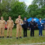 Grupa osób w mundurach beżowych i czarnych stoi na trawie. W tle są zielone drzewa i niebieskie oraz zielone namioty. Jedna osoba robi zdjęcie telefonem.