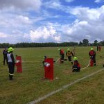 Grupa strażaków w zielonych kaskach ćwiczy na zielonym polu pod pochmurnym niebem. Tło to drzewa i czerwony wóz strażacki. Widać czerwone przeszkody i białe węże gaśnicze.