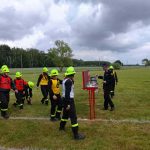 Na polu widoczni są strażacy w neonowych kaskach i kolorowych kamizelkach. Tło stanowi pochmurne niebo i drzewa. Strażacy zdają się uczestniczyć w ćwiczeniach lub zawodach.