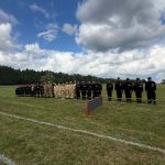 Grupa strażaków w czarnych i beżowych mundurach stoi na zielonej łące. Niebo jest niebieskie z białymi chmurami. Obok nich znajduje się czerwony stół z nagrodami.