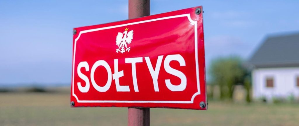 Czerwona tablica z napisem SOŁTYS i białym orłem. W tle widoczne pole i fragment budynku. Dominują kolory czerwony i niebieski.