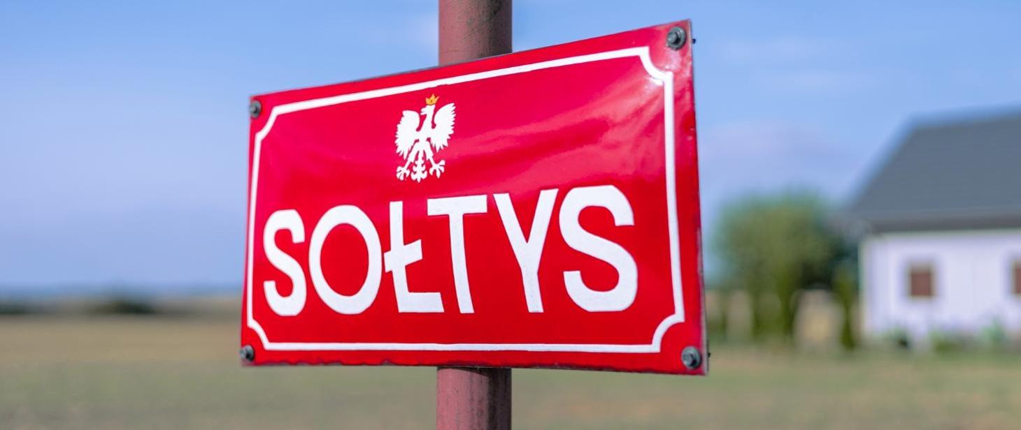 Czerwona tablica z napisem SOŁTYS i białym orłem. W tle widoczne pole i fragment budynku. Dominują kolory czerwony i niebieski.