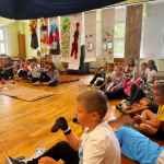 4 Grupa dzieci siedzi na podłodze w jasnej sali z drewnianą podłogą. W tle wiszą kolorowe kostiumy oraz prace plastyczne. Dzieci wydają się uczestniczyć w warsztatach lub zajęciach kreatywnych.