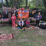 Grupa dzieci z instrumentami, w parku z zielonym tłem, przy pomarańczowym banerze. Kolorowe stroje i instrumenty. Akustyczna gitara i śpiew znakują wydarzenie.