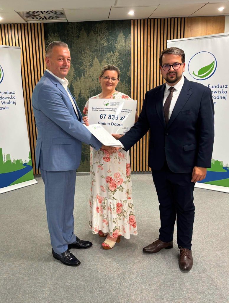 Trzy osoby w eleganckich strojach trzymają czek na tle drewnianej ściany i banerów. Kobieta w kwiecistej sukience. Kolory stonowane z zielonym akcentem.