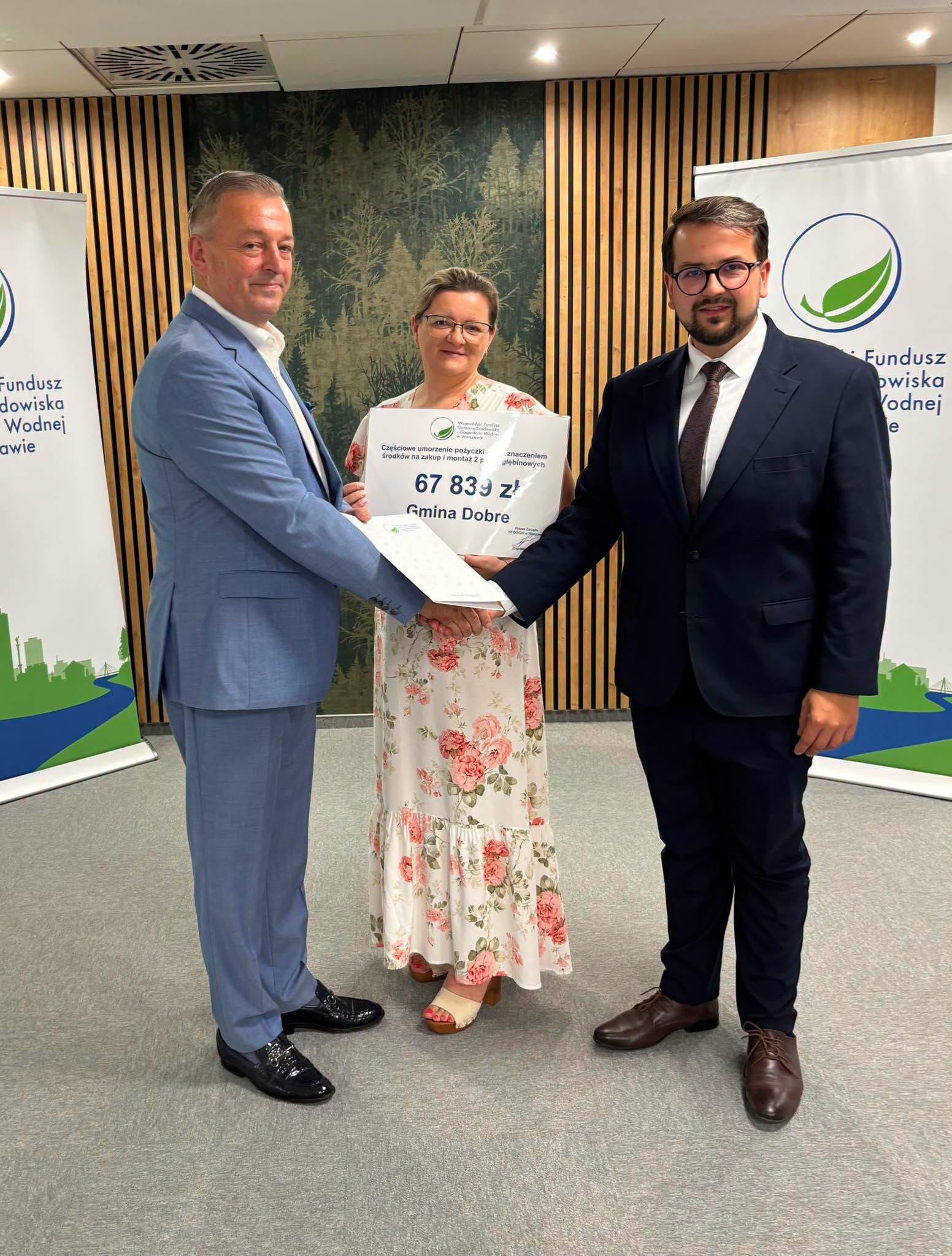 Trzy osoby w eleganckich strojach trzymają czek na tle drewnianej ściany i banerów. Kobieta w kwiecistej sukience. Kolory stonowane z zielonym akcentem.