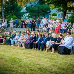 Na zdjęciu widzimy dużą grupę ludzi siedzących na krzesłach, ubranych kolorowo, na tle zieleni i domów. Wydarzenie odbywa się na zewnątrz, widoczne są drzewa i trawnik. Atmosfera jest uroczysta, a ludzie uważnie obserwują wydarzenie.