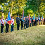 Grupa strażaków w mundurach stoi z flagami na tle zielonego lasu. Pojawiają się czerwone i niebieskie barwy flag. Tło to trawiaste podłoże i drzewa.