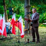 Mężczyzna w garniturze przemawia na zewnątrz z mikrofonem w tle drzew. Obok powiewają polskie flagi. W tle zieleń i zgromadzeni ludzie.
