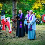 Na zdjęciu trzej duchowni w fioletowych szatach stoją na trawie, wokół biało-czerwone flagi. Tło to zielone drzewa, a w oddali widać czerwony wóz strażacki. Wydarzenie odbywa się na świeżym powietrzu.