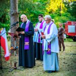 Trzej duchowni w fioletowych stułach stoją na trawie przy mikrofonie, na tle lasu. Obok polska flaga, w tle wóz strażacki. Ubrania w kolorach czerni i bieli z fioletowymi akcentami.