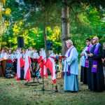 Grupa osób stoi na trawie, w tym duchowni w strojach liturgicznych. W tle zespół muzyczny z białymi i czerwonymi flagami. Otoczenie to zielone drzewa.