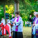 Kapłani w białych szatach i fioletowych stułach stoją przy mikrofonie na tle drzew. Obok biało-czerwone flagi. Tło to zielona sceneria zebranych ludzi.