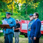Na zdjęciu trzech mężczyzn w mundurach, w lesistym otoczeniu. Jeden mówi przez mikrofon, w tle czerwony wóz strażacki. Dominują zielenie i czerwienie.