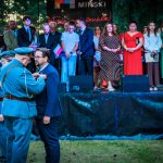 Na scenie odbywa się uroczystość. Osoba w mundurze przypina medal mężczyźnie. W tle stoi grupa ludzi w kolorowych strojach.