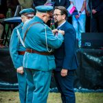 Mężczyzna w niebieskim mundurze przypina odznaczenie do marynarki innego. Tło z grupą obserwujących osób, niektórzy trzymają flagi Polski. Kolory dominujące: niebieski i zielony.