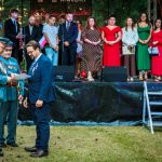 Grupa ludzi na scenie z zielonym tłem. Dwie osoby w mundurach wręczają dokumenty, ludzie w eleganckiej odzieży. Chłopiec w bieli podchodzi po schodach.