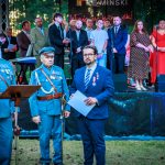 Grupa osób na scenie; dwie w mundurach, jedna w garniturze. W tle zielone drzewa. Ubiór w głównie niebieskich i czerwonych barwach.