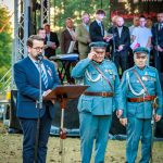 Trzy osoby na scenie, w tym dwóch mężczyzn w niebieskich mundurach wojskowych. Mężczyzna w garniturze przemawia do mikrofonu. W tle grupa ludzi i zielone drzewa.