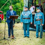 Mężczyzna w garniturze przemawia na tle mikrofonu, obok dwóch mundurowych z medalami. Tło to zielone drzewa i scena.