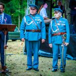 Trzech mężczyzn podczas uroczystości na świeżym powietrzu. Dwaj w mundurach wojskowych z medalami, trzeci w garniturze przy mikrofonie. Tło zielone, leśne.