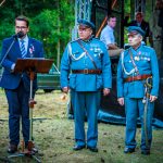 Mężczyzna w garniturze przemawia przy mikrofonie. Obok niego stoją dwaj starsi panowie w niebieskich mundurach z medalami. Tło to zalesiony teren i scena z różnymi osobami.