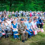 Grupa ludzi siedzi na zewnątrz, wiele z nich trzyma polskie flagi. W tle zielone drzewa i tłum stojący. Dzień jest słoneczny.