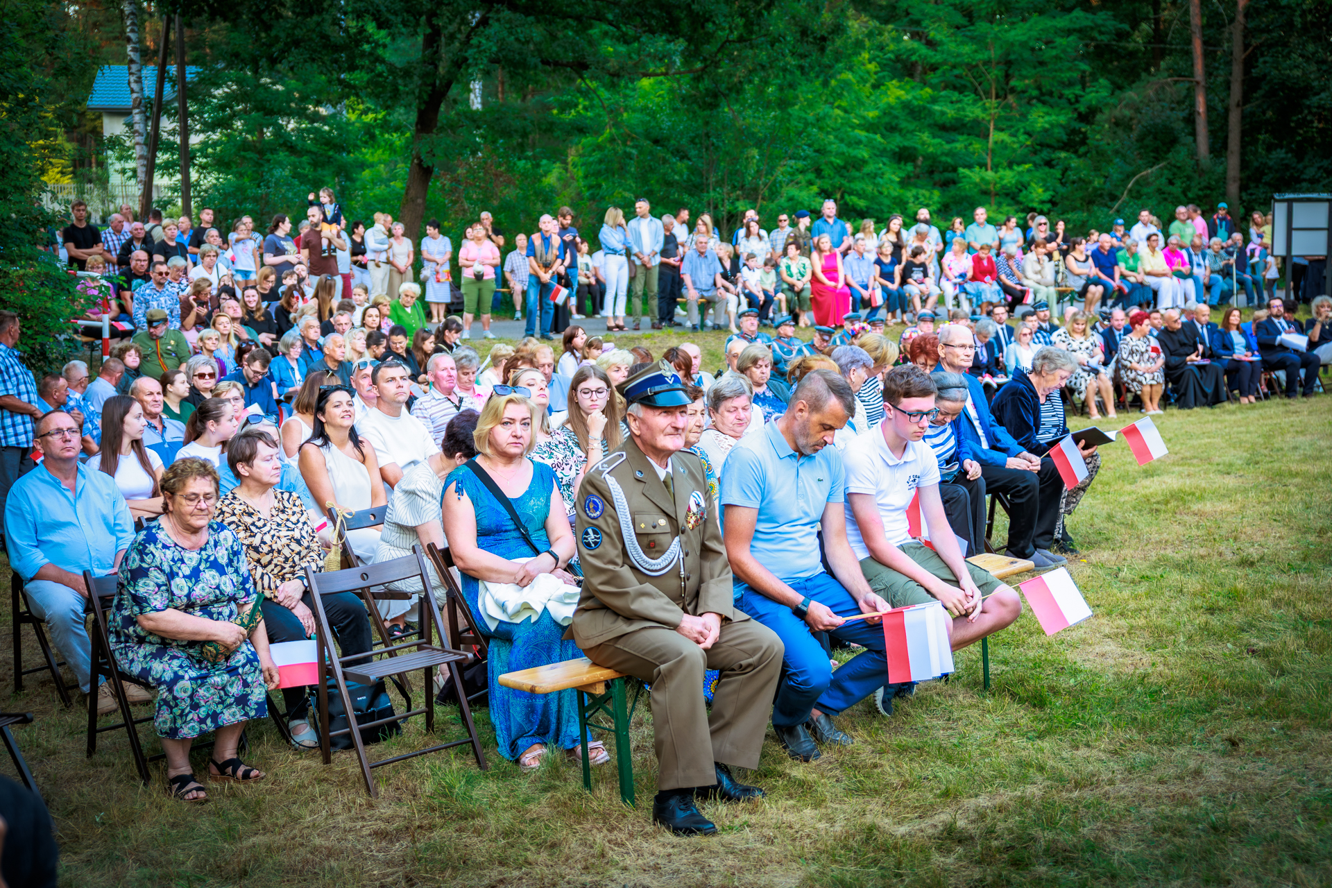 Grupa ludzi siedzi na zewnątrz, wiele z nich trzyma polskie flagi. W tle zielone drzewa i tłum stojący. Dzień jest słoneczny.