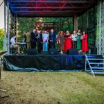 Grupa ludzi śpiewa na scenie plenerowej, otoczonej zielenią drzew. Tło sceny jest czarne z napisem „Powiat Miński”. Osoba z mikrofonem stoi z boku, a muzyk gra na klawiszach.