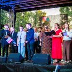 Grupa ludzi śpiewa na scenie w plenerze. Kolorowe ubrania, w tle zielone drzewa. Pianista po lewej stronie.