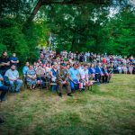 Na zdjęciu widoczna jest zgromadzona na trawie grupa ludzi z polskimi flagami. Tło stanowią zielone drzewa. Ubrania uczestników są w różnych kolorach.