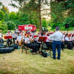 Orkiestra dęta występuje na świeżym powietrzu, otoczona zielenią. Muzycy ubrani są w białe koszule i czarne spodnie. W tle czerwona ciężarówka strażacka.