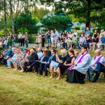 Grupa osób siedzi na krzesłach na trawniku. W tle drzewa i budynek, kolorystyka zielono-niebieska. Atmosfera przypomina wydarzenie plenerowe.