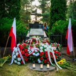 Grób z kwiatami w kolorze czerwonym i białym, otoczony polskimi flagami. W tle zielone drzewa i krzyż. Osoba w niebieskiej odzieży stoi przy grobie z zapalonym zniczem.