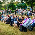 Grupa ludzi siedzi na krzesłach na trawie przy leśnej scenerii, w pierwszym rzędzie duchowni w fioletowych szatach.